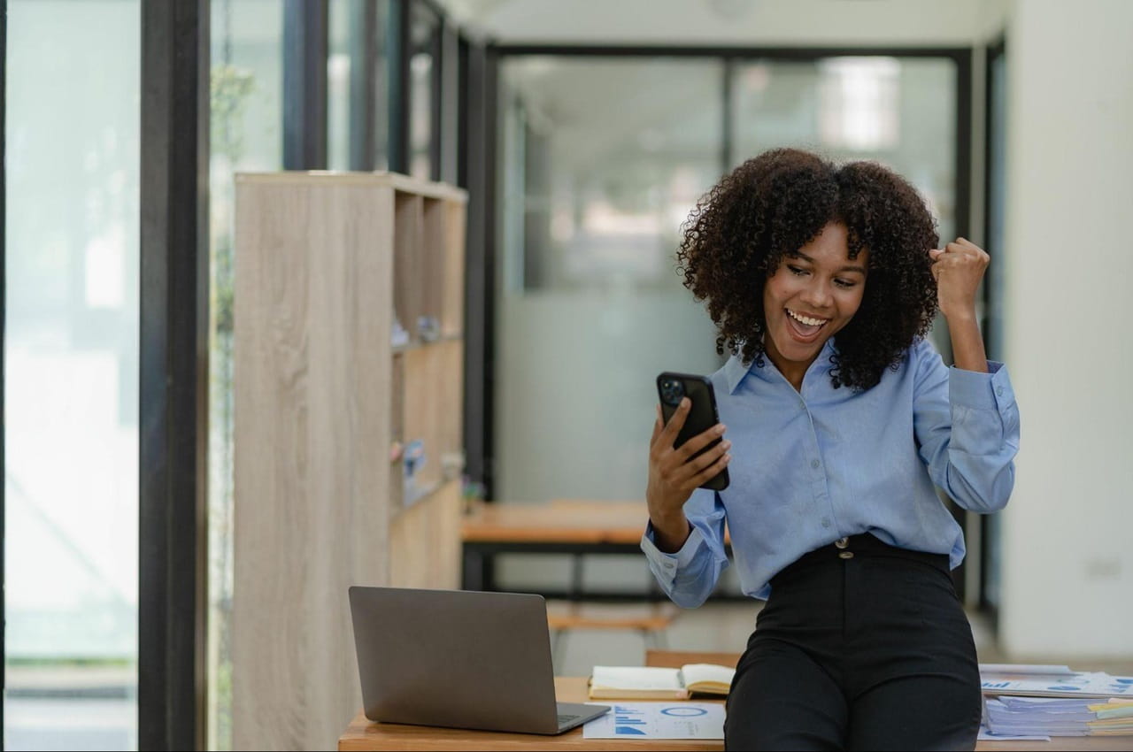 Mulher sorridente veste camisa e calça social e se apoia em mesa de escritório, enquanto comemora olhando para o celular.