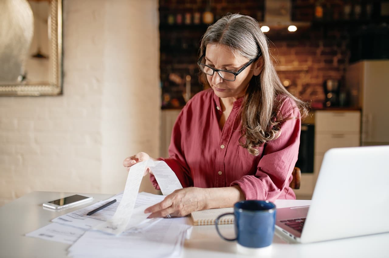 Mulher sênior com expressão concentrada veste blusa social vermelha e analisa extratos e holerites ao lado de um laptop em sua residência.