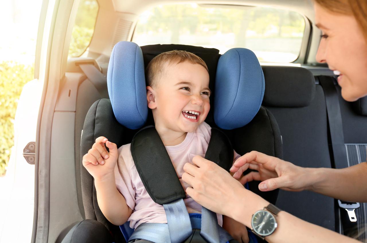 Mulher sorridente colando cinto em um bebê de colo sorridente que está na cadeirinha de proteção de um carro.