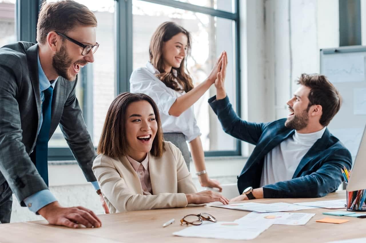 Grupo de pessoas sorridentes sentadas ao redor de uma mesa de escritório com papéis em cima. As pessoas vestem casacos e ternos de alfaiataria. 