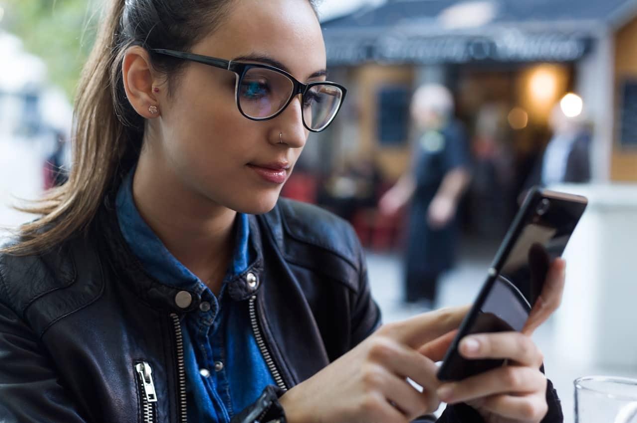 Mulher de óculos de grau e jaqueta de couro preta digitando no celular sobre golpes de empréstimo. Ela está sentada em uma praça de alimentação.