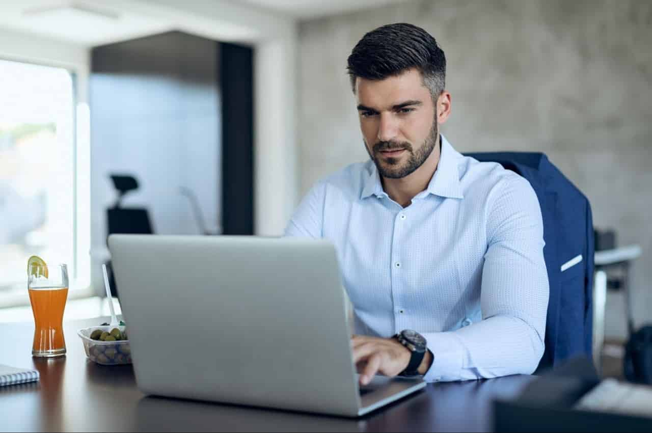 Homem de expressão concentrada, vestindo blusa social azul e digitando no computador da empresa.