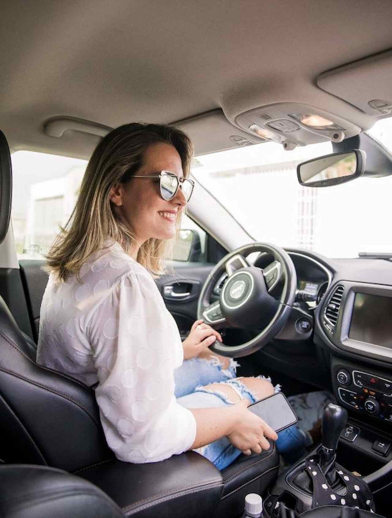 A imagem mostra uma mulher loira, usando óculos escuros e dirigindo seu carro.