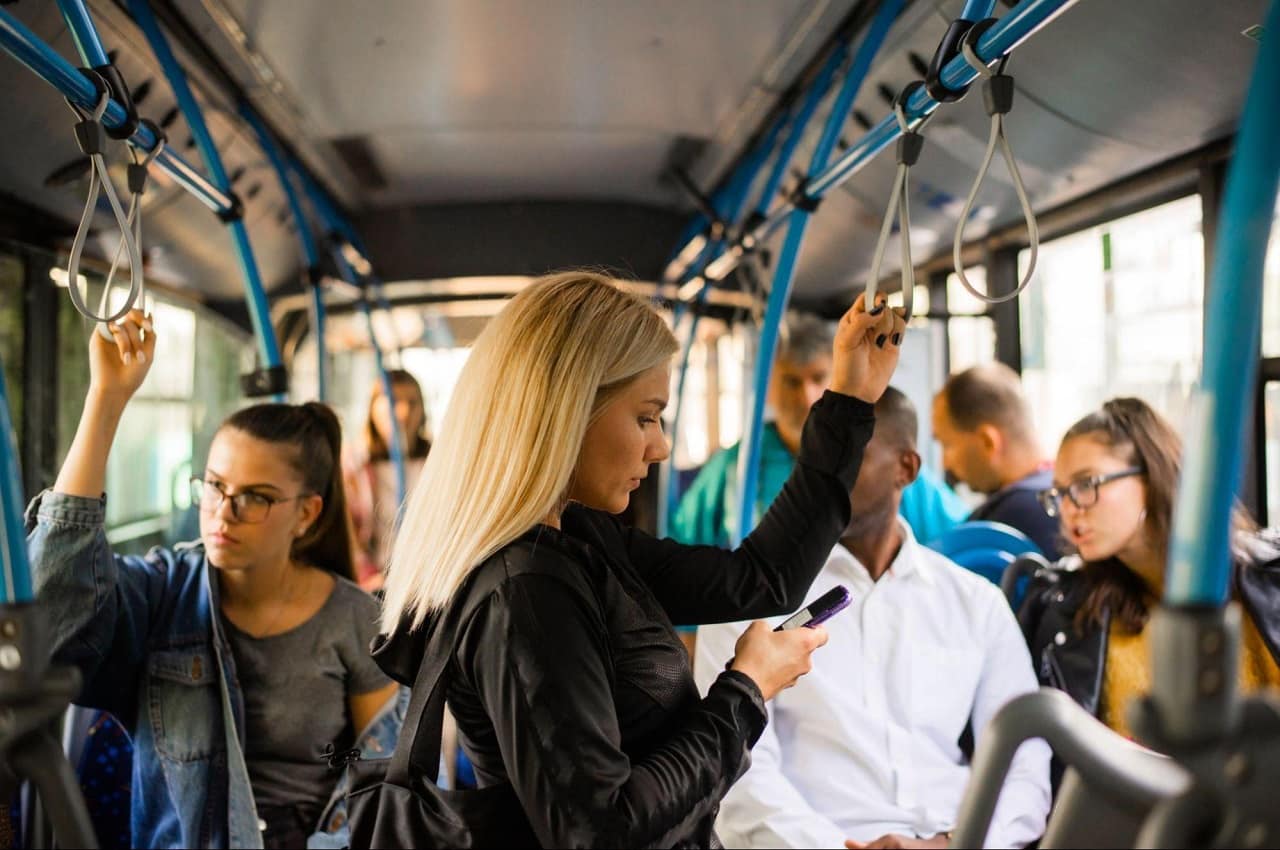 Pessoas em pé dentro do transporte público (ônibus) e foco em uma mulher loira digitando no celular.