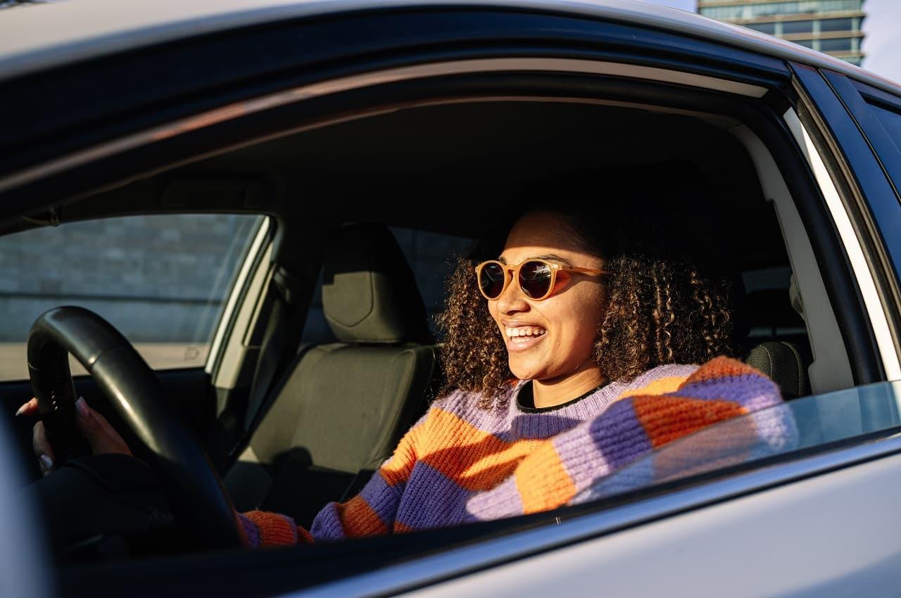 Mulher negra de óculos escuros e blusa listrada laranja e roxa, com expressão sorridente e dirigindo um carro.