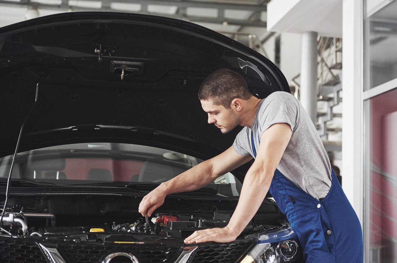 Mecânico com macacão de segurança azul, segurando chave de fenda e fazendo manutenção veicular no carro para família.