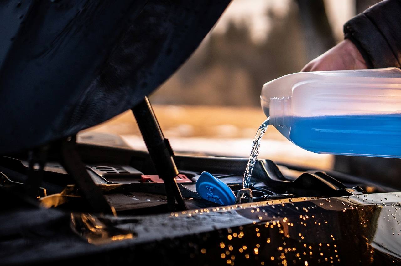 Mecânico (seu rosto não aparece na imagem) jogando líquido azul no motor de um carro próprio para realizar a manutenção veicular.