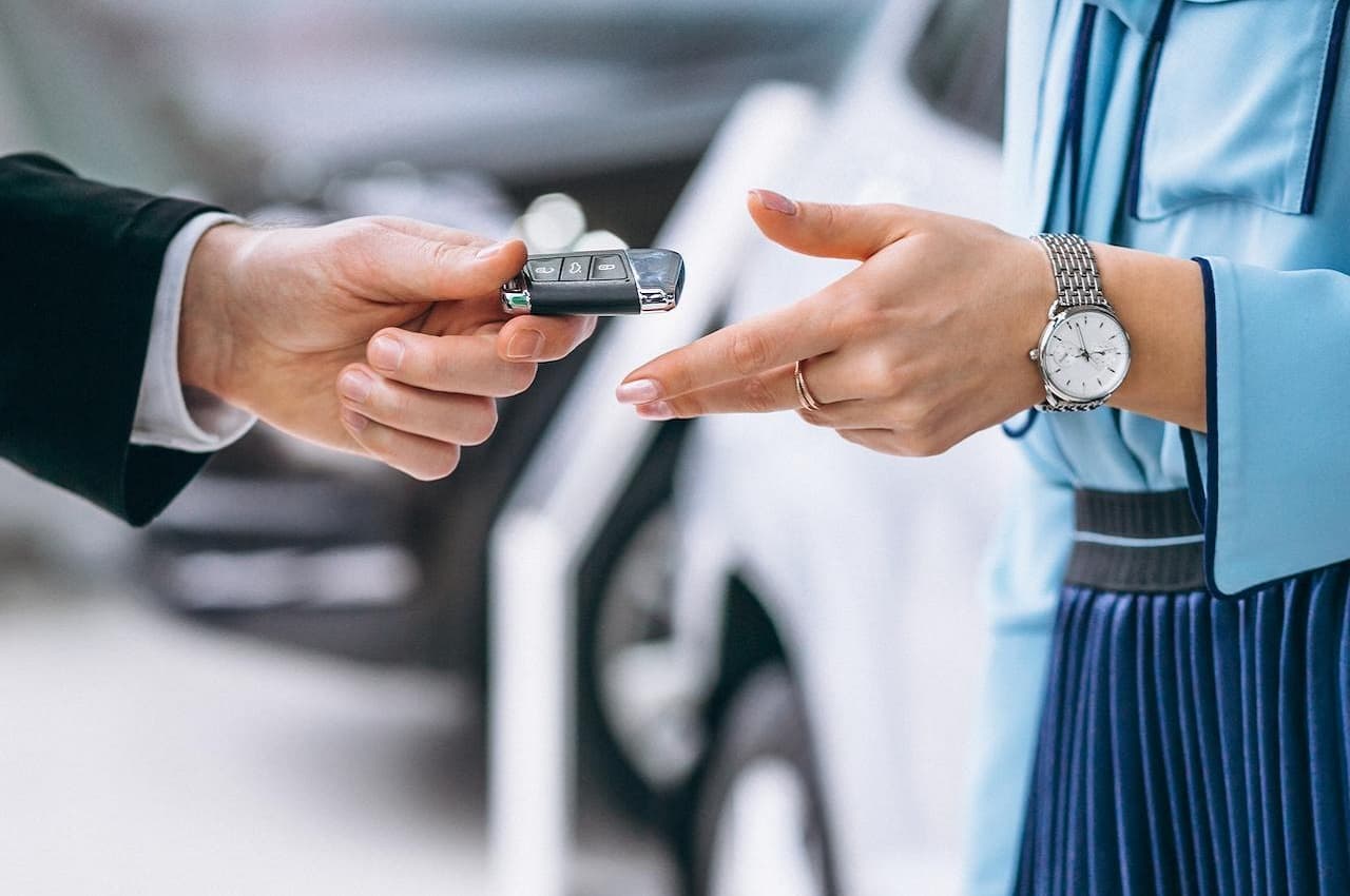 Foco na mão de um homem entregando chaves de carro para uma mulher. Os rostos das pessoas não aparecem na imagem. O homem veste terno preto e a mulher camisa social listrada e calça jeans.