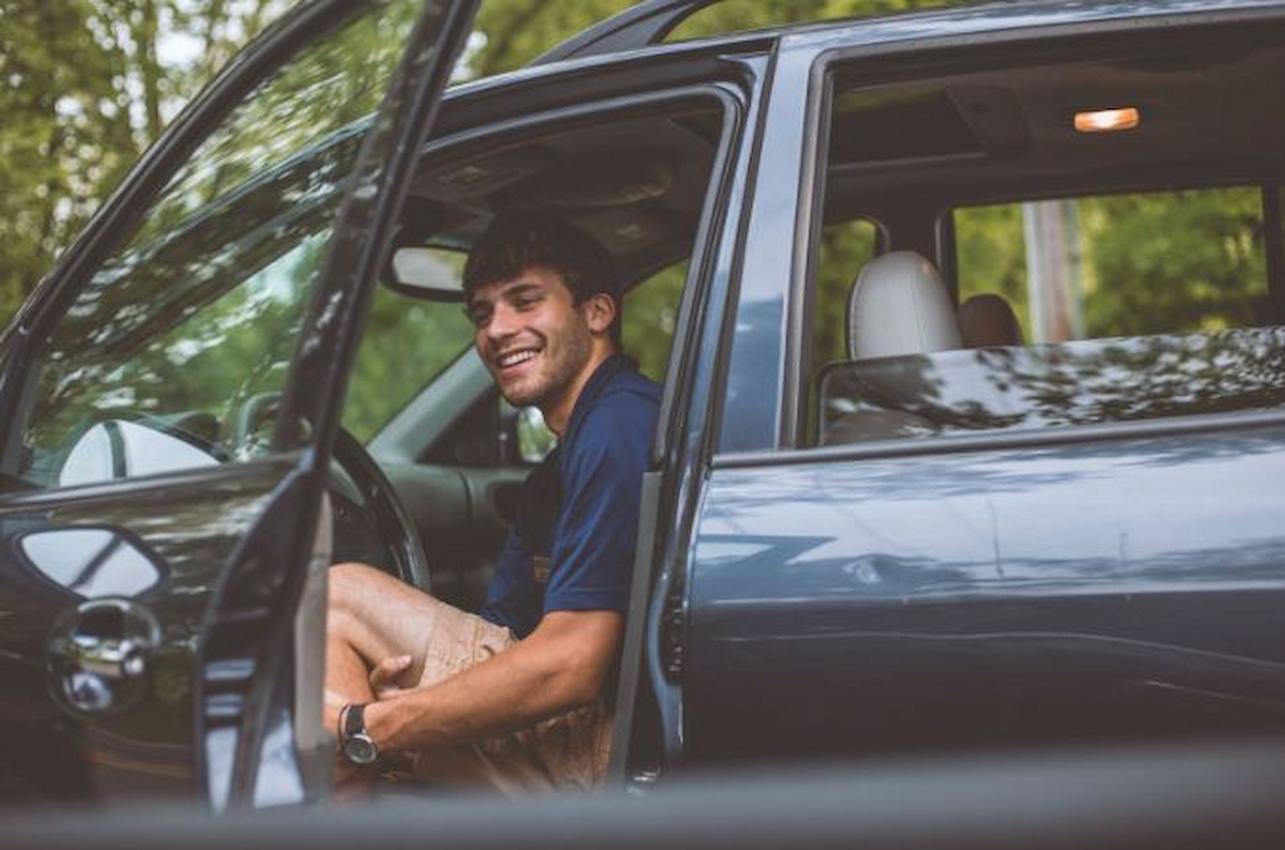 A imagem mostra um rapaz sentado ao banco do motorista de um carro. A porta está aberta.