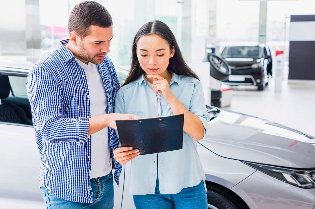 A imagem mostra um homem e uma mulher analisando um papel em uma concessionária, próximos a um carro.