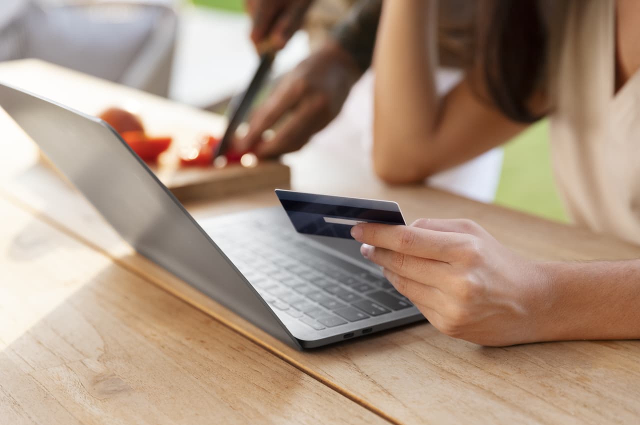 A imagem mostra uma pessoa usando seu laptop e com seu cartão de crédito em uma mão. Ao seu lado, outra pessoa cortando tomates.