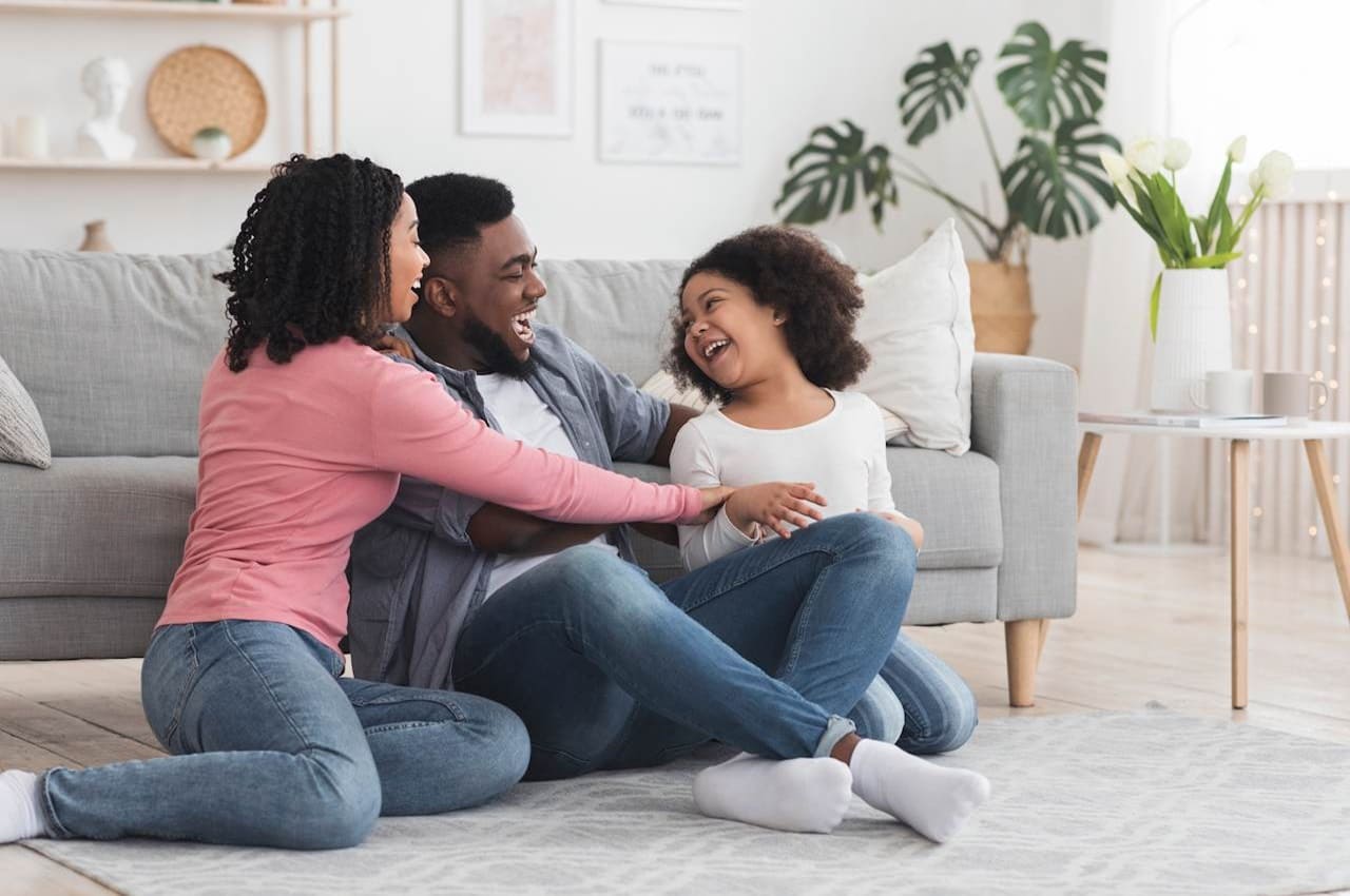 Família sorridente (homem, mulher e criança) sentados no tapete de sua casa e abraçados.