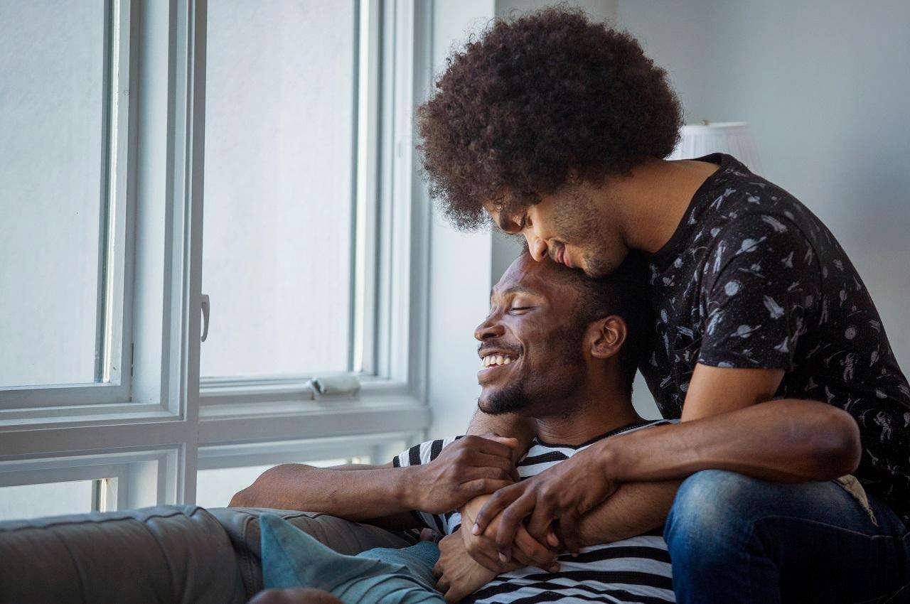 Casal sorridente composto por dois homens que se abraçam e observam a paisagem por uma janela fechada.