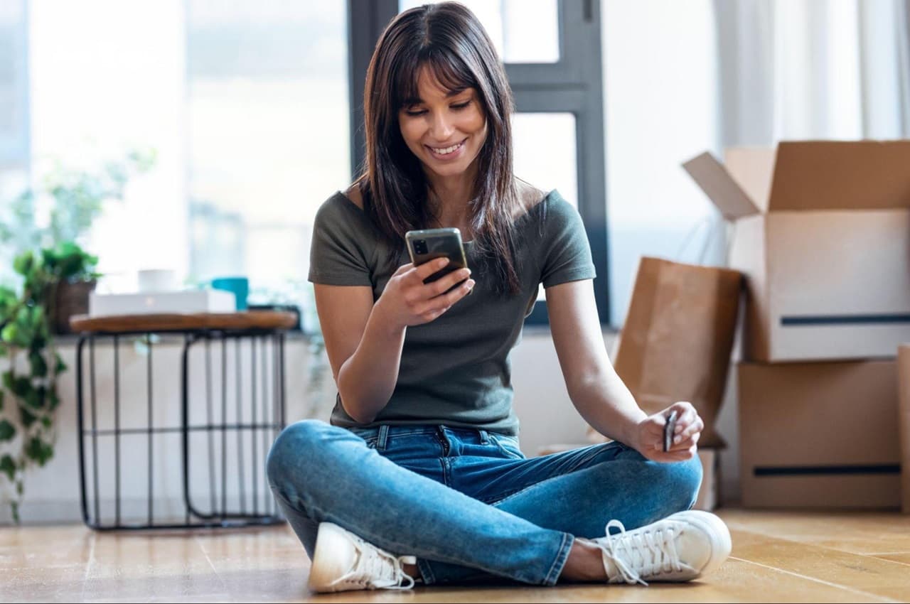 Mulher sorridente de cabelos lisos castanhas e franja, sentada no chão de casa digitando no celular. A mulher veste blusa cinza básica sem mangas e calça jeans.