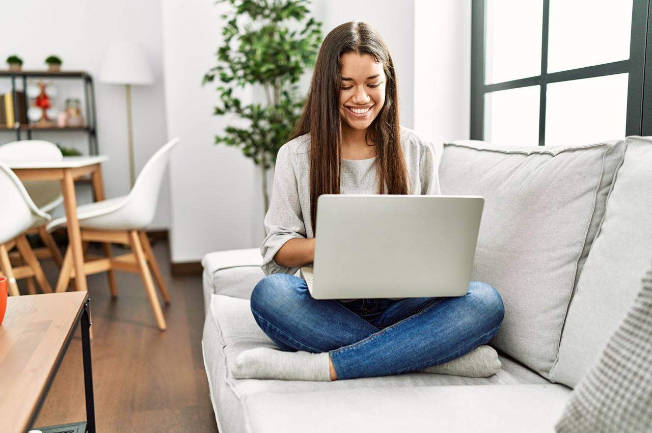 Mulher sorridente de cabelos castanhos longos, sentada de perna cruzada no sofá e segurando laptop no colo.