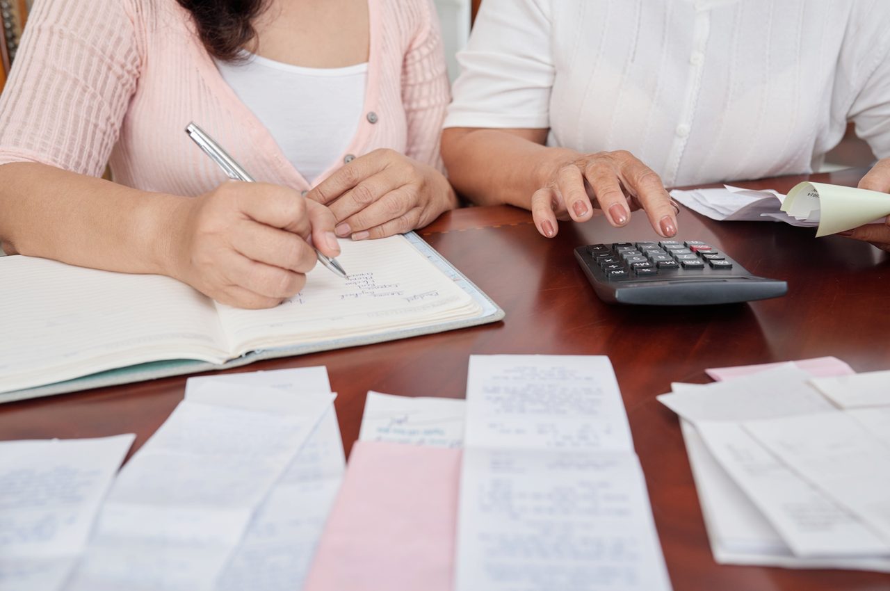 A imagem mostra duas mulheres fazendo as contas do mês em uma calculadora.