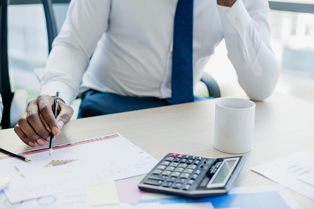 Homem com camiseta social branca e gravata azul, calculando taxa pré e pós fixada na calculadora e analisando gráfico de barras no escritório.