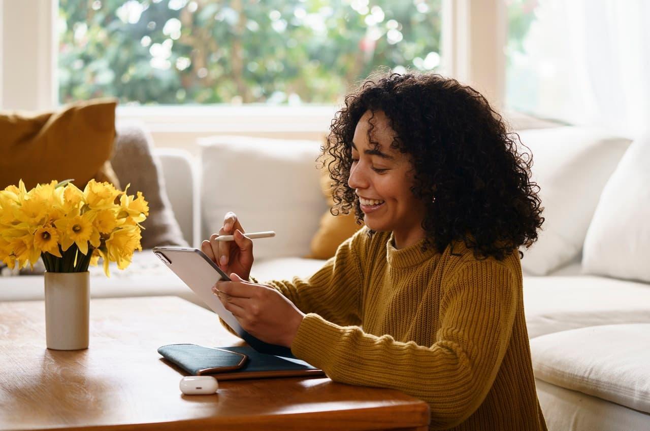 Mulher de cabelo crespo sentada à mesa e sorrindo enquanto usa um tablet. Ela está pesquisando sobre a parcela de financiamento de veículos.