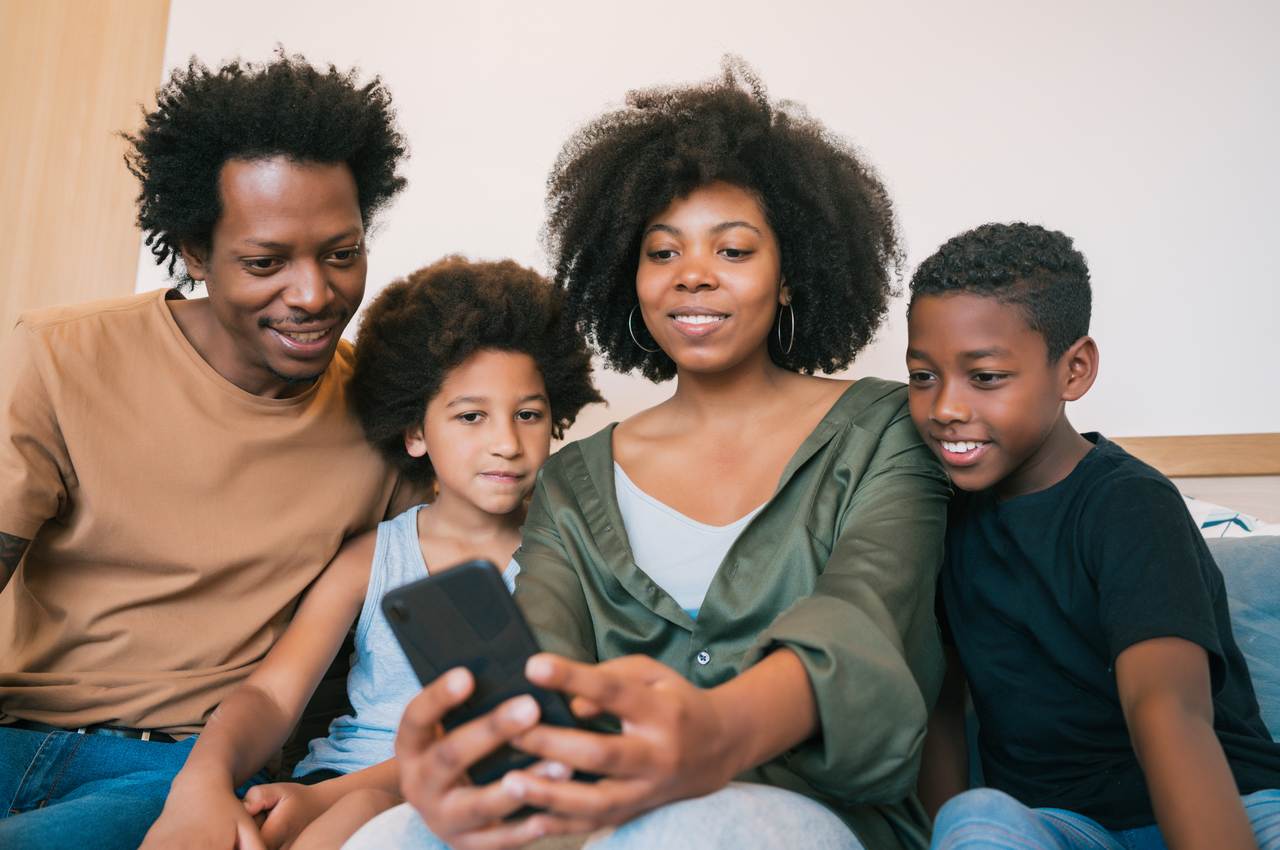 Família sorridente composta por um homem, uma mulher e uma criança. Todos olham para a tela de um celular.