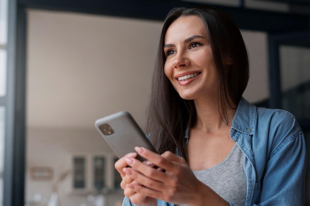 Mulher sorridente de cabelos castanhos longo, utilizando celular para acessar Shopping BV. A mulher veste camisa jeans e regata cinza.