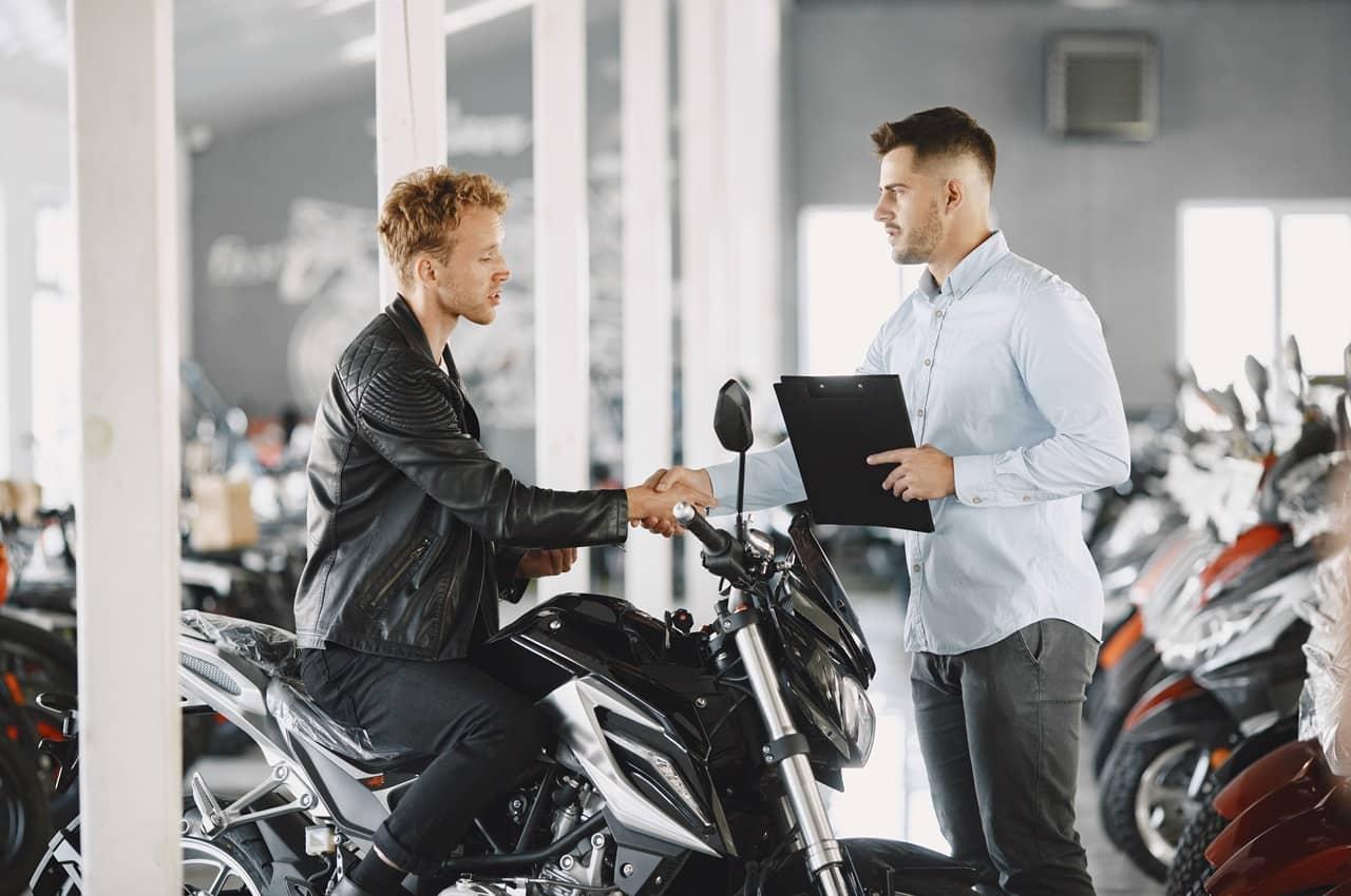 Homem comprando primeira moto em uma concessionária. Ele está fazendo um sinal de aperto de mãos com o dono da loja.