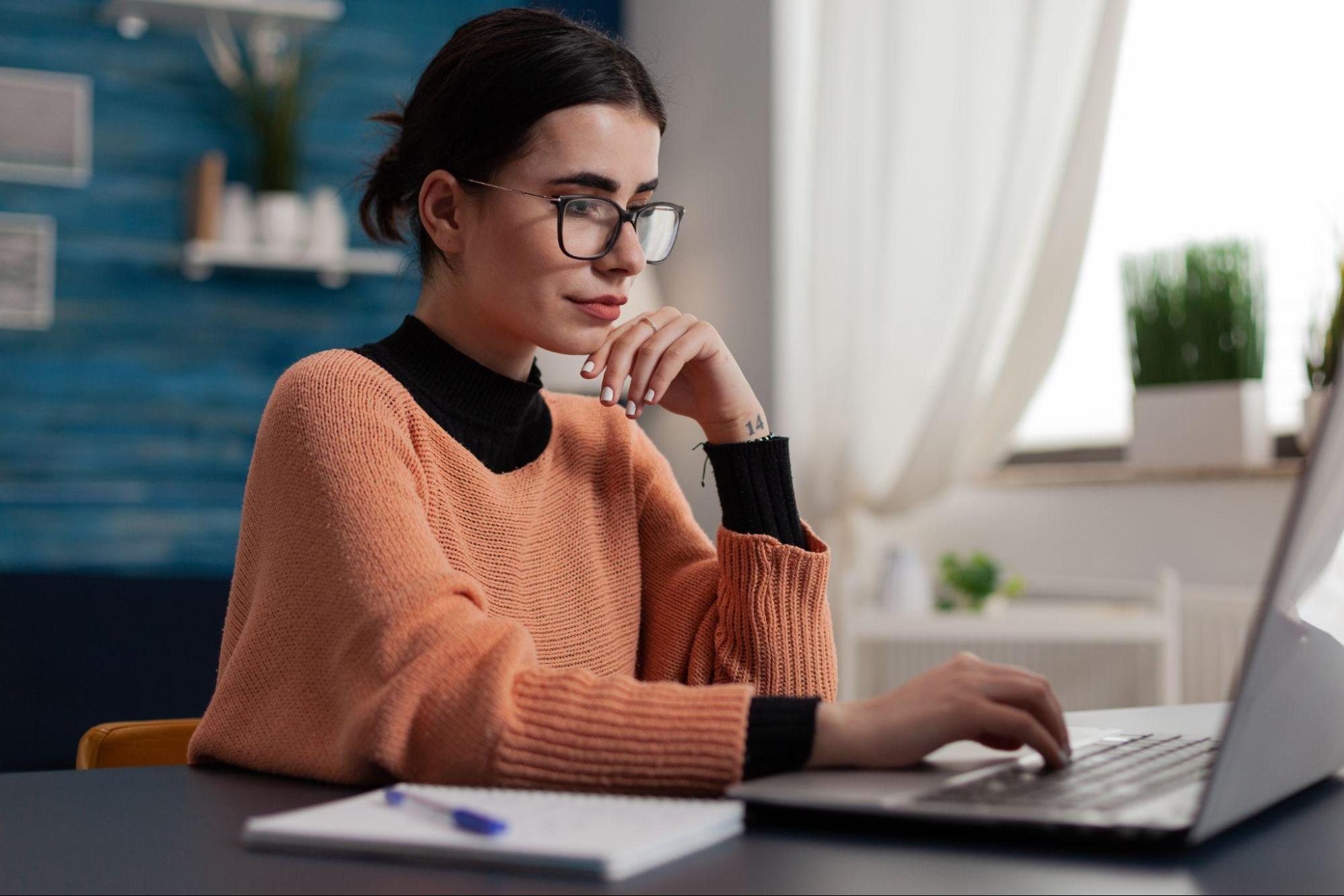 Mulher com expressão concentrada e cabelo preso em coque, usa um laptop aberto em sua mesa do escritório e acessa empréstimo para CLT. A mulher veste blusa de tricot laranja e usa óculos de grau.