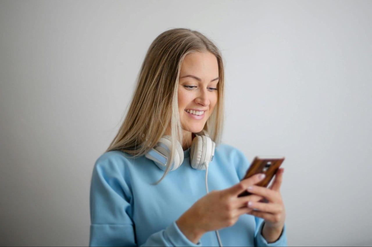 Mulher loira sorridente usa fones ouvido sem fio, suéter azul claro e acessa empréstimo pessoal BV pelo celular.