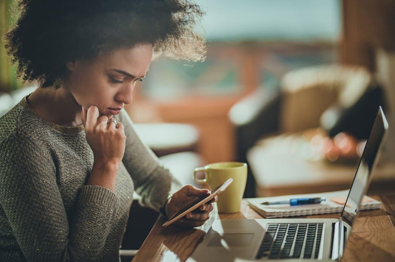 Mulher com expressão concentrada consultando gravame pelo celular e usando laptop em cima de uma mesa.