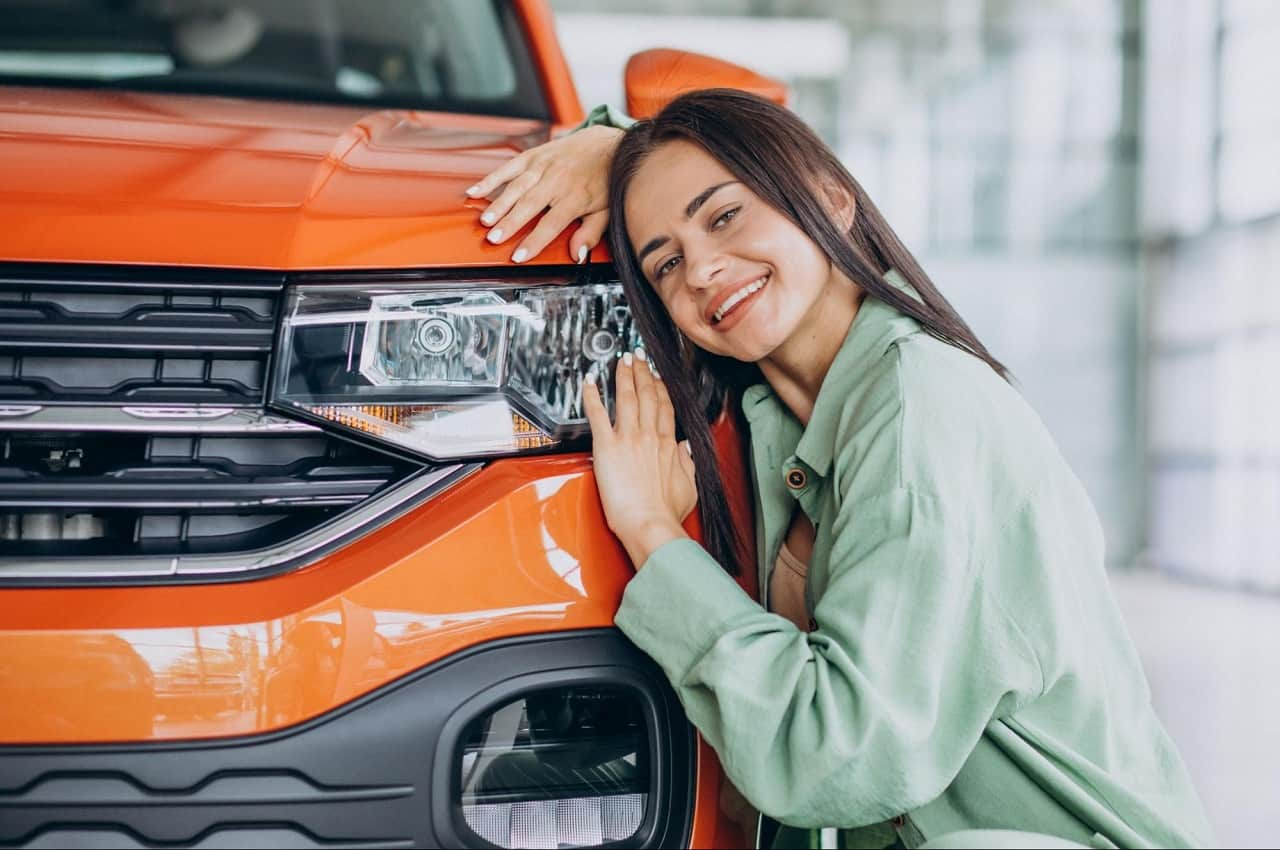 A imagem mostra uma mulher encostada nos faróis de um carro laranja.