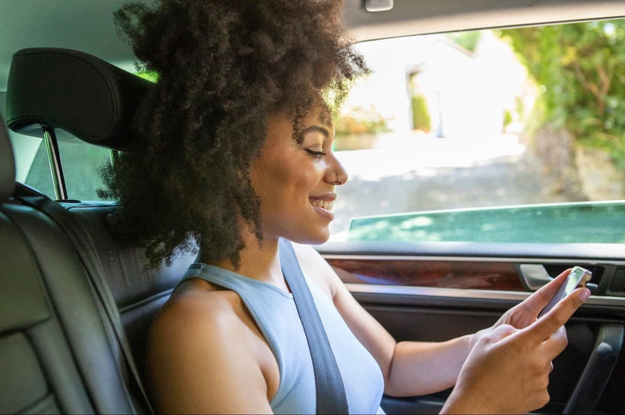 Mulher negra sorridente, sentada no banco de motorista do carro estacionado e digitando no celular.