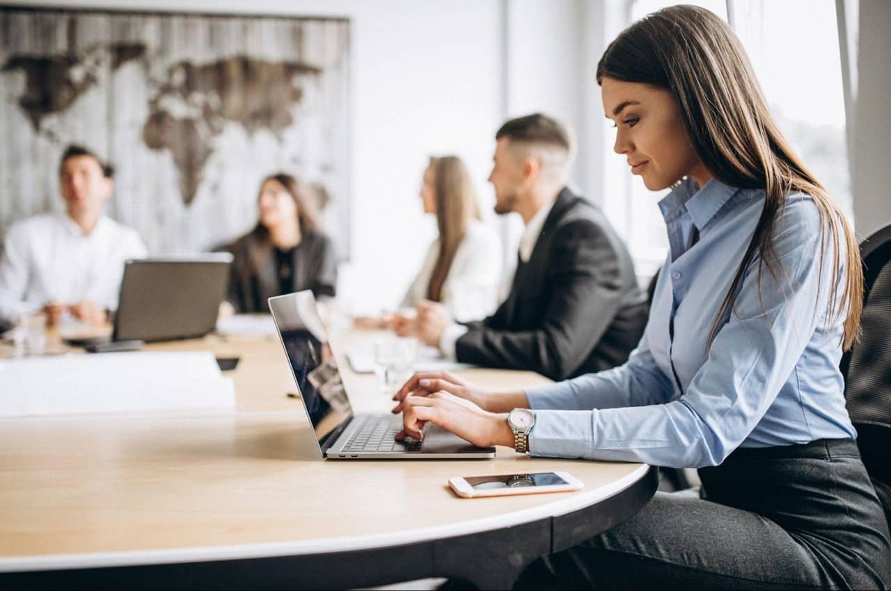 Grupo de pessoas sorridentes no trabalho em volta de uma mesa. A imagem foca em uma mulher sentada digitando no computador da empresa.