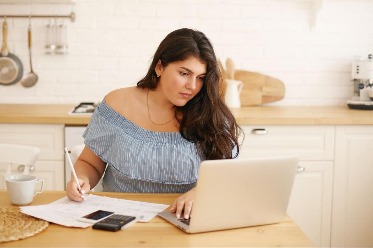 Mulher com expressão concentrada, olhando para tela de um computador e anotando no papel informações de como declarar carro financiado no IR.