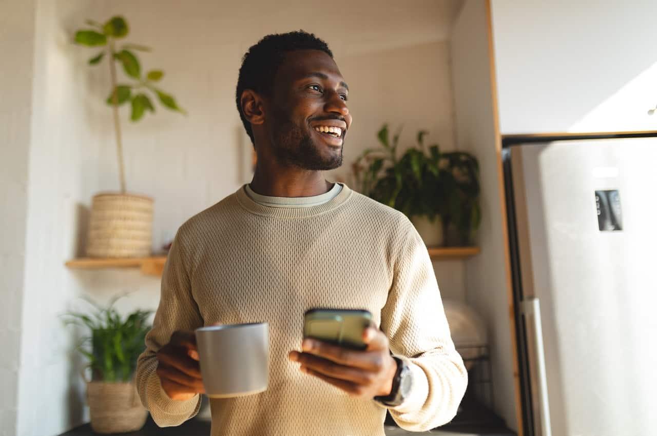 Homem sorridente, vestindo blusa de linho e em pé na cozinha de casa, olhando para o lado e segurando um celular e uma xícara.