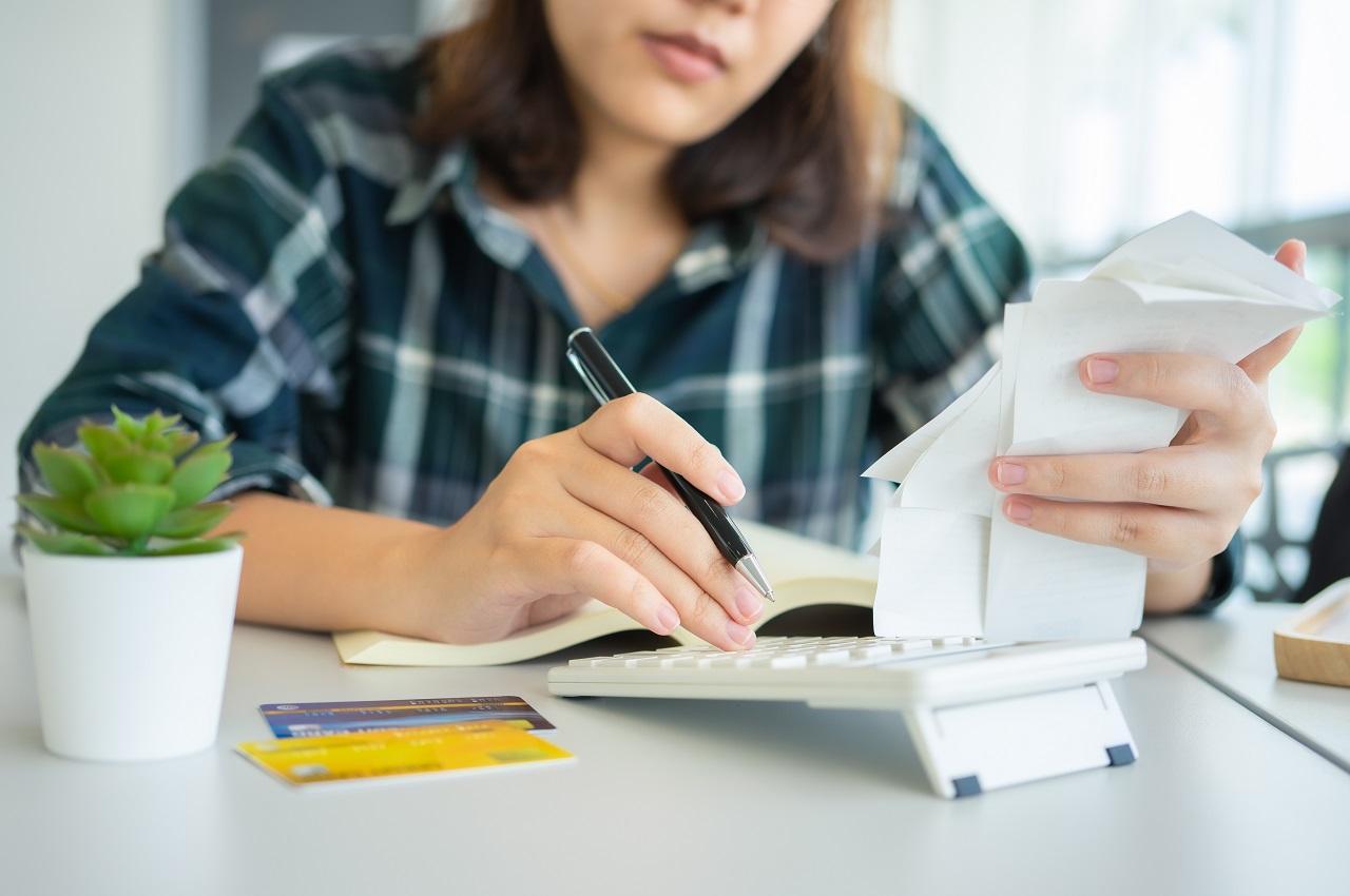 Mulher de cabelos castanhos curtos, utilizando calculadora e um computador para anotar suas compras por impulso.