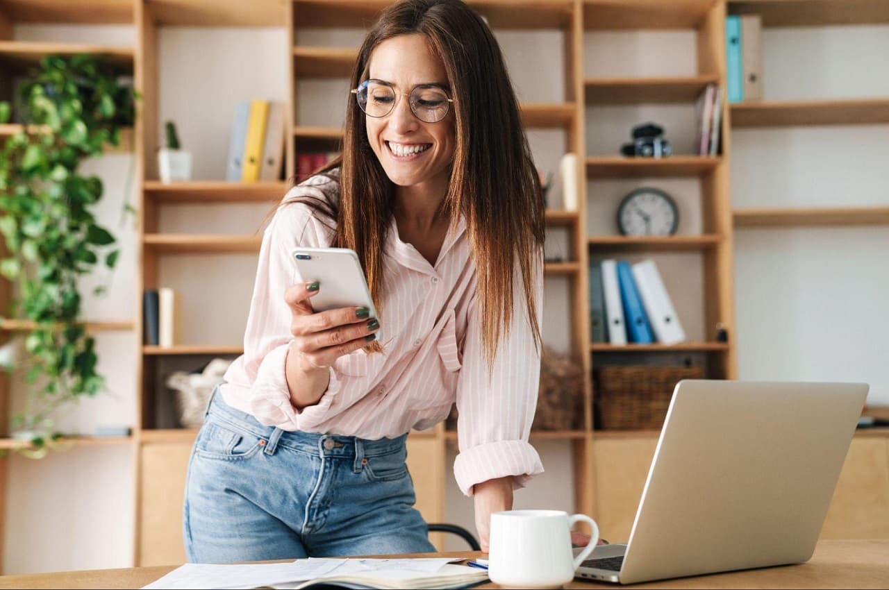 Mulher sorridente de cabelos castanhos médios e óculos de grau, utilizando celular e apoiando sua mão na mesa do escritório. Ela veste calça jeans e camisa social listrada.