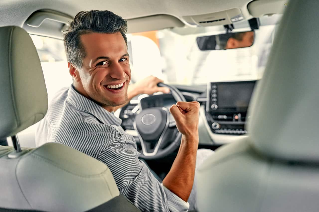 Homem sorridente olhando para trás, fazendo um gesto de vitória após ter financiado carro sem entrada. Ele se encontra sentado no banco do motorista.