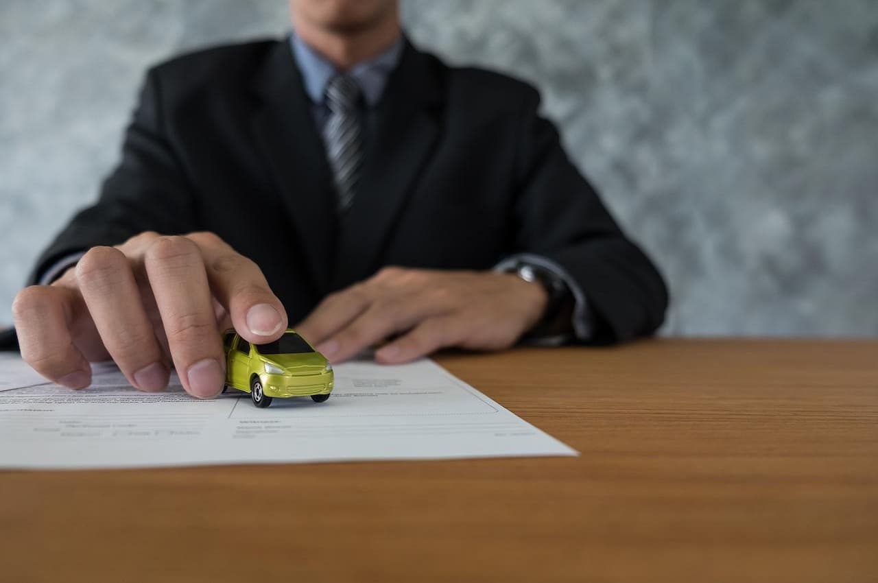 Homem de terno no escritório (seu rosto não aparece na imagem), segurando miniatura de carro amarelo de quatro portas em cima de uma mesa de madeira.