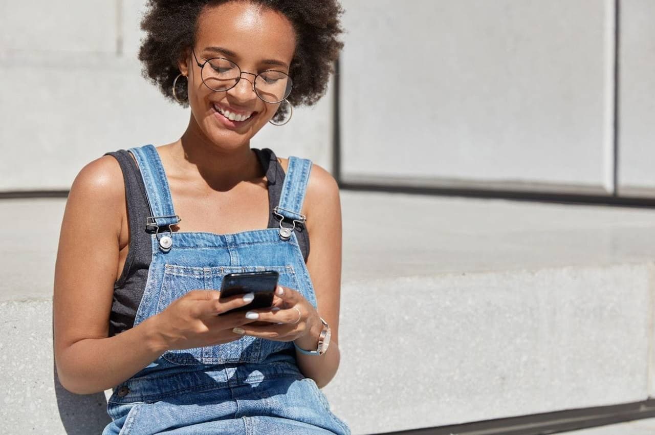 Mulher sorridente com óculos de grau, apoiada na parede de um estabelecimento e acessando portabilidade de salário pelo celular. A mulher veste macacão jeans e camisa básica preta.