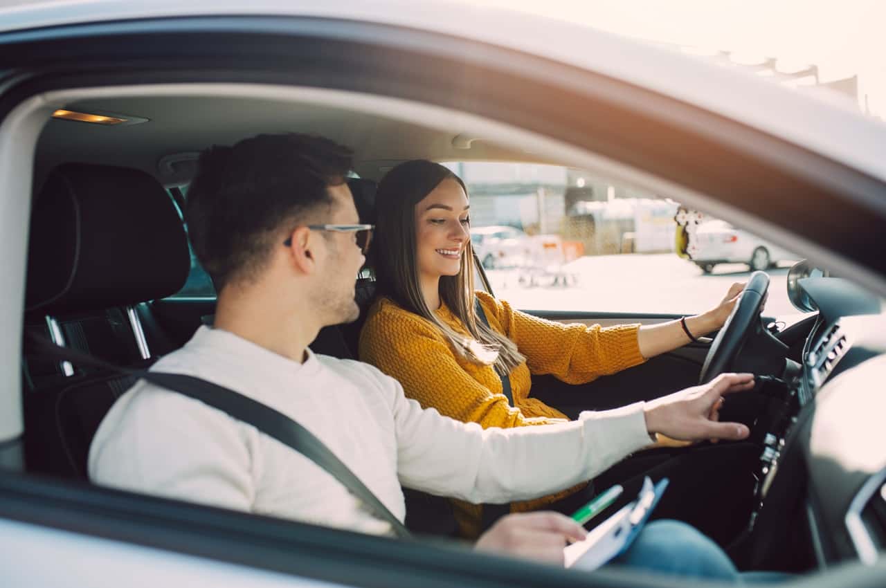 Duas pessoas dentro de um carro a mulher de cabelos compridos e blusa amarela esta na direção, enquanto um homem de perfil conversa apontando para o painel do veiculo