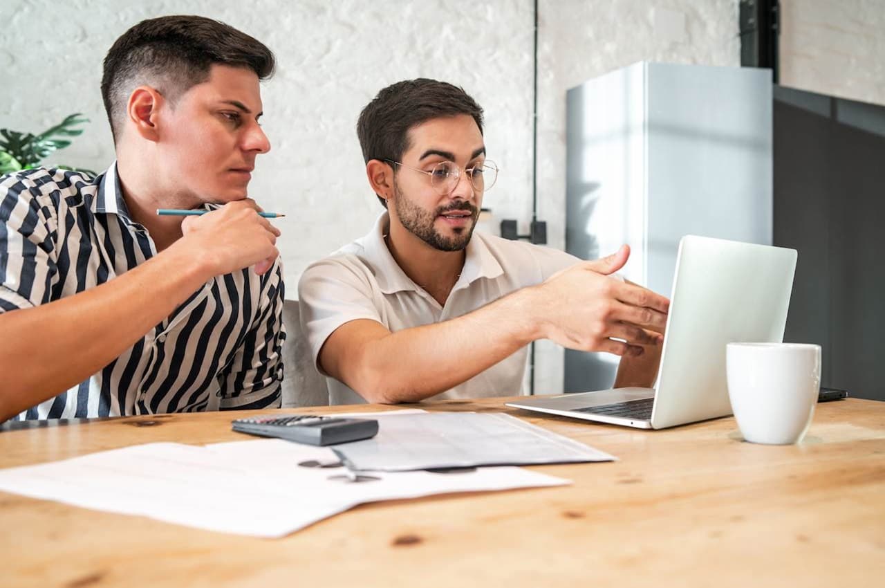 Dois homens conversando entre si no escritório. Um deles aponta para a tela de um laptop e o outro mantém uma expressão concentrada.