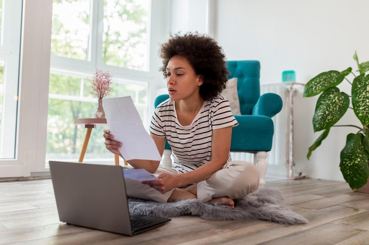 Mulher negra com expressão concentrada lendo uma folha de papel e utilizando computador para conseguir empréstimo consignado. Ela está sentada no tapete da sala,veste camisa listrada preta e branca e calça moletom.