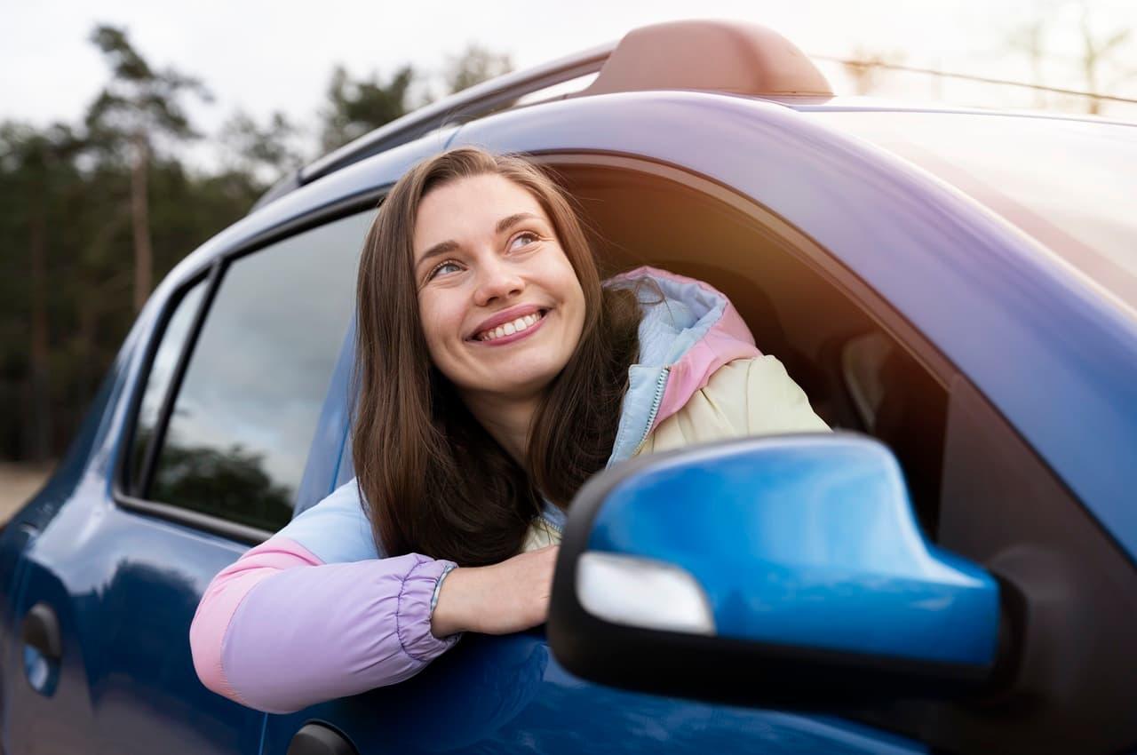 Mulher sorridente de cabelos castanhos lisos, apoiada na janela de um veículo azul estacionado.