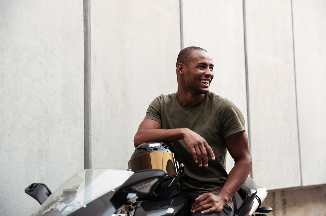 A imagem mostra um rapaz, usando uma camiseta verde-musgo em cima de sua moto.