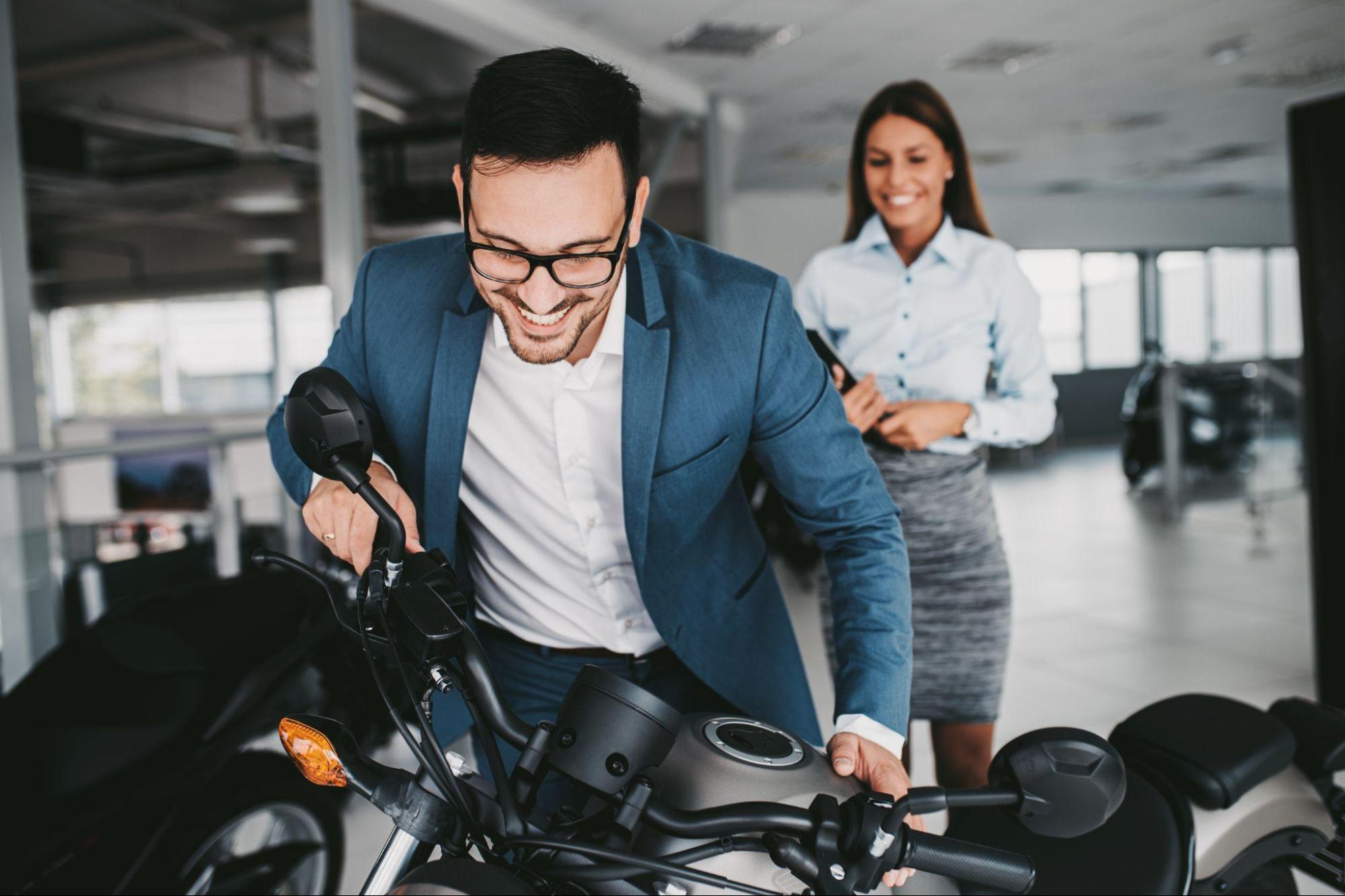 Homem sorridente em terno azul examinando uma motocicleta, enquanto uma mulher ao fundo, também sorridente, o observa.