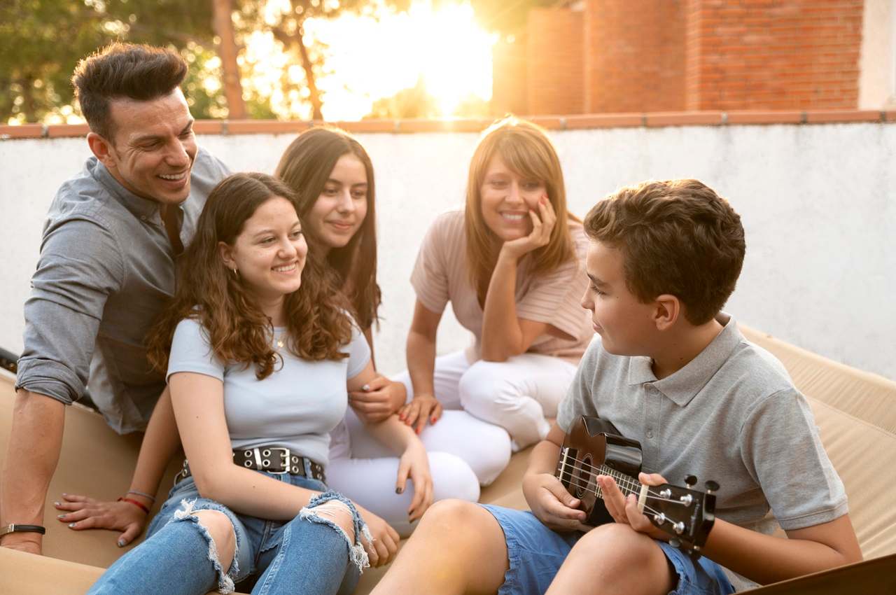 A imagem mostra uma família sorridente e reunida. Um garoto está tocando violão. 