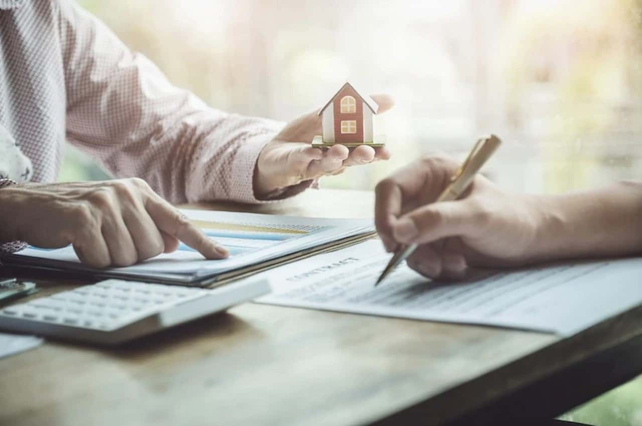 Pessoa (rosto não aparece na imagem) assinando contrato de seguro residencial, enquanto o vendedor segura uma miniatura de uma casa nas mãos.