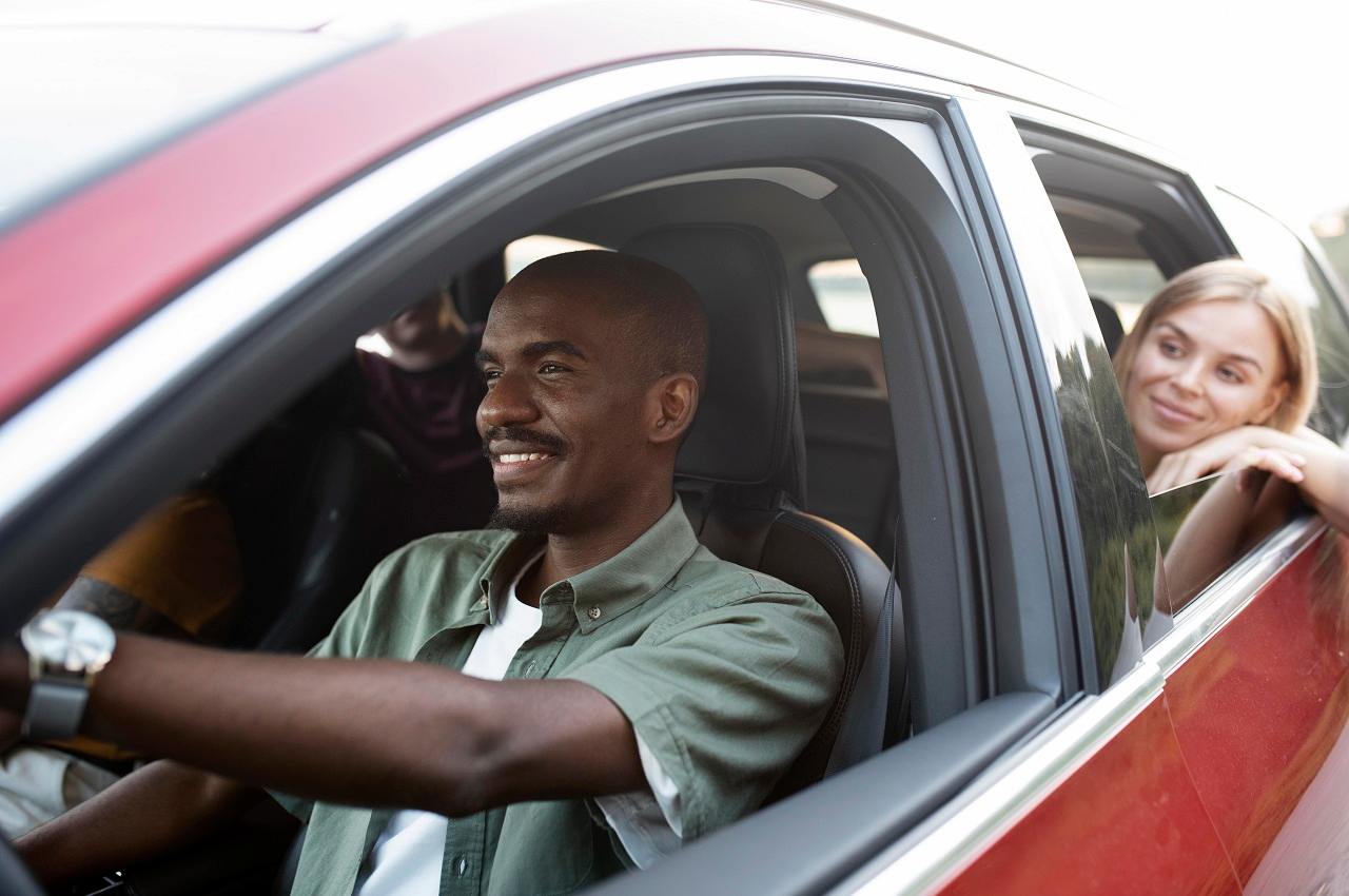 Homem sorridente dirigindo carro vermelho no test drive e levando três passageiros (um homem e duas mulheres).