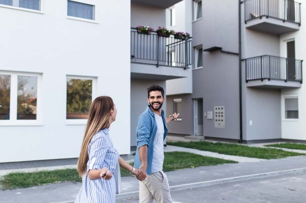 Homem sorridente segurando a mão de uma mulher sorridente e apontando para um apartamento a venda.