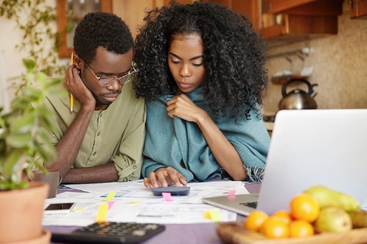 Casal com expressão concentrada utilizando calculadora e computador para verificar seguro renda protegida. O homem veste blusa social verde e a mulher um chale azul.