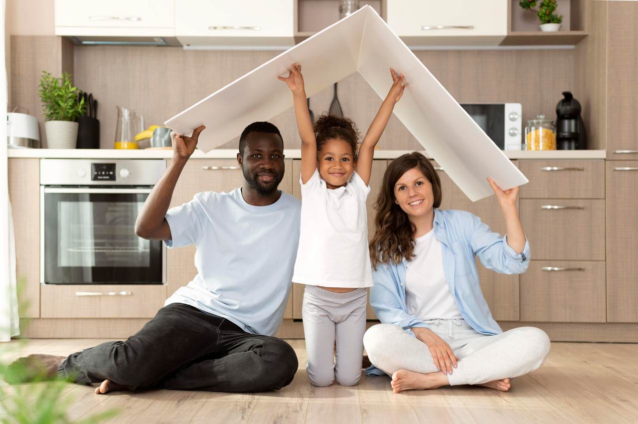 Família sorridente (homem, mulher e uma criança) segurando papelão em cima da casa para simular telhado de uma casa. Todos estão sentados no chão de sua nova residência.